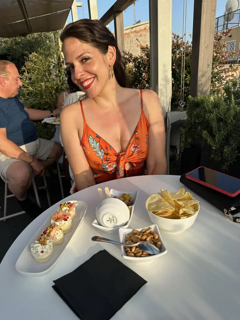 Lindsay Taylor Dellinger sitting at a table outside, smiling shyly at camer with head tilted to one side - aperitivi on the table