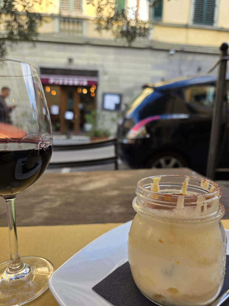 glass of wine and jar of tiramisu at forefront; storefront and car in background